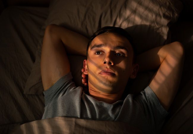 Молодой мужчина в серой футболке лежит в постели с руками за головой
